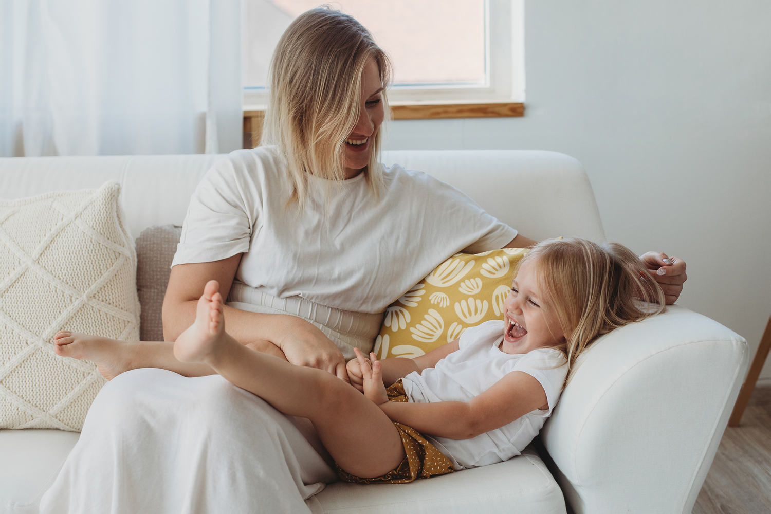 Mother and toddler daughter sitting on a couch with pillows and laughing together