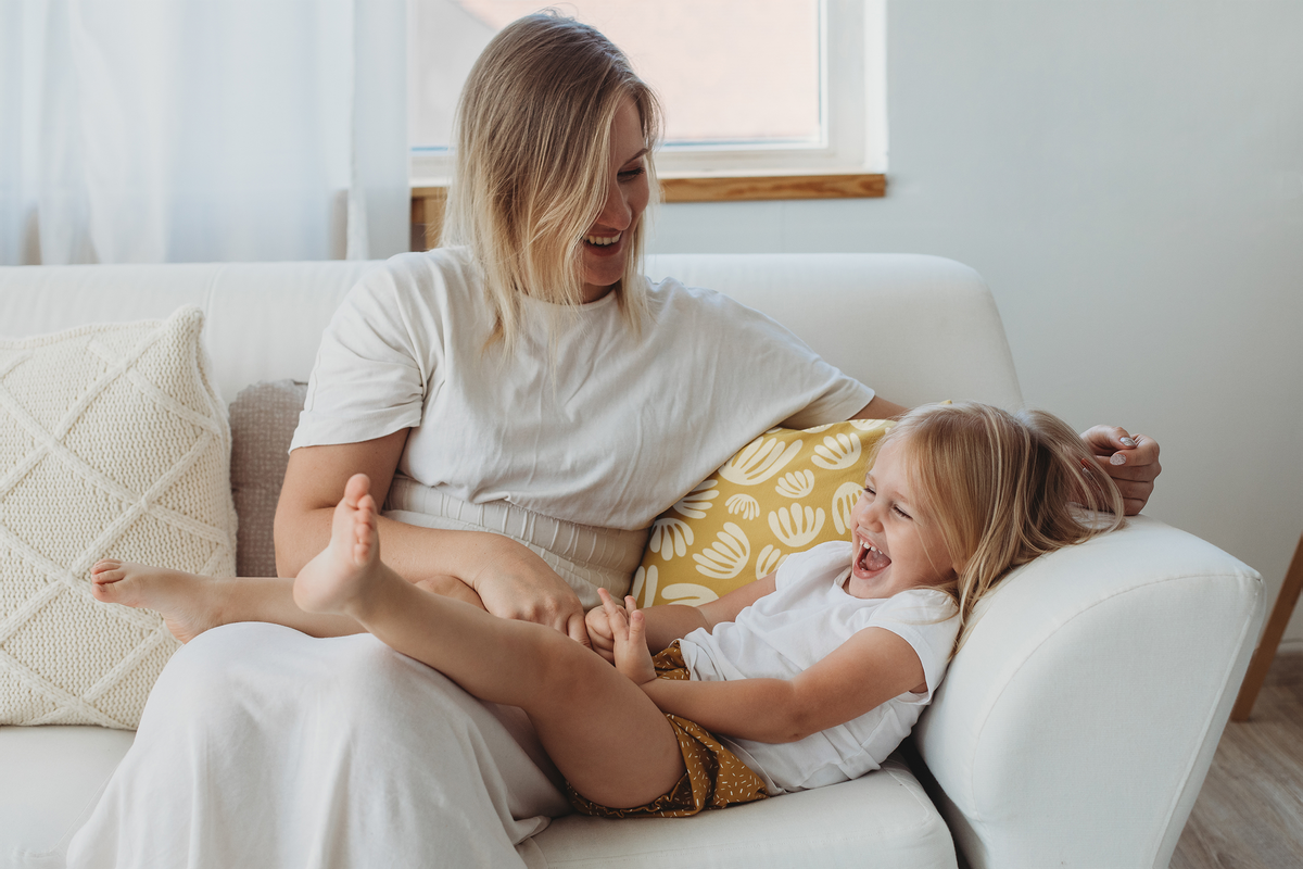 Mother and toddler daughter sitting on a couch with pillows and laughing together